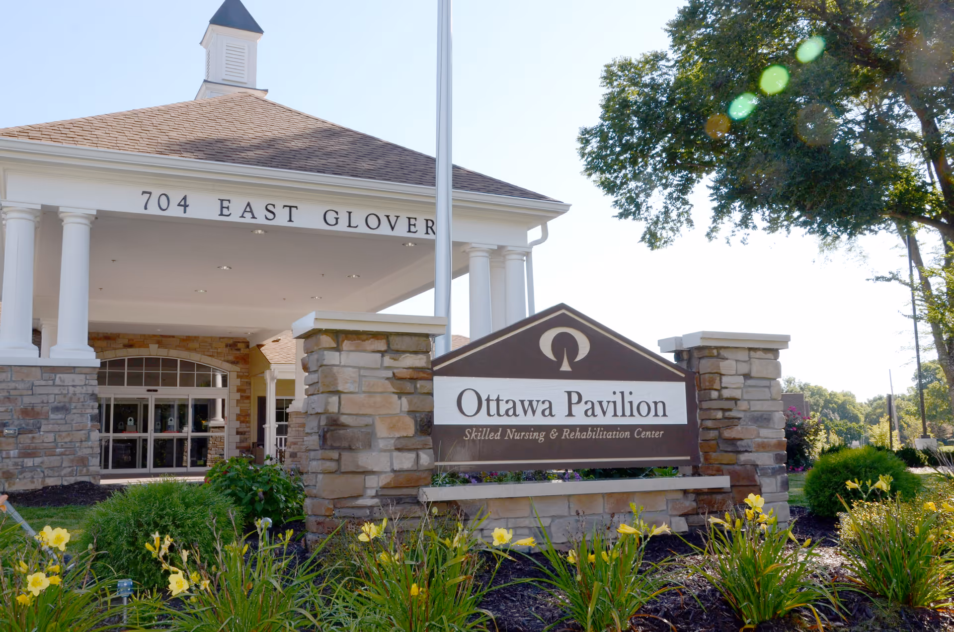 Exterior view of Ottawa Pavilion Skilled Nursing & Rehabilitation Center with a stone sign in front surrounded by yellow flowers and greenery. The building entrance has white columns and a peaked roof with the address 704 East Glover visible.