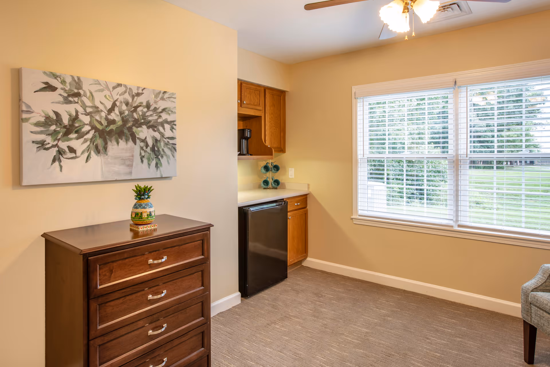 Interior view of a room with beige walls and carpeted floor. A wooden chest of drawers with a decorative pineapple on top is positioned against one wall beneath a painting of green leaves. To the right, there is a small kitchenette area with wooden cabinets, a black mini fridge, and a coffee maker. A large window with white blinds lets in natural light and shows a green outdoor area. A ceiling light fixture with multiple bulbs is visible above.