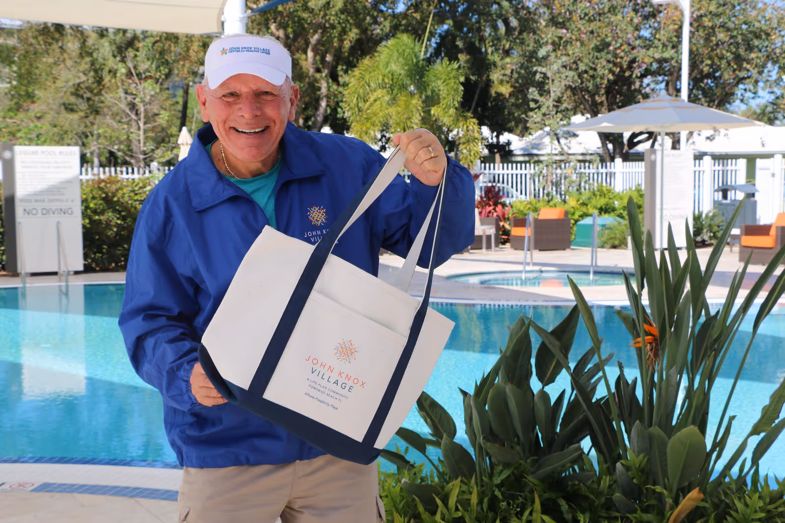 An elderly man wearing a blue John Knox Village jacket and white cap is smiling and holding a John Knox Village tote bag near a swimming pool with greenery and outdoor seating in the background.