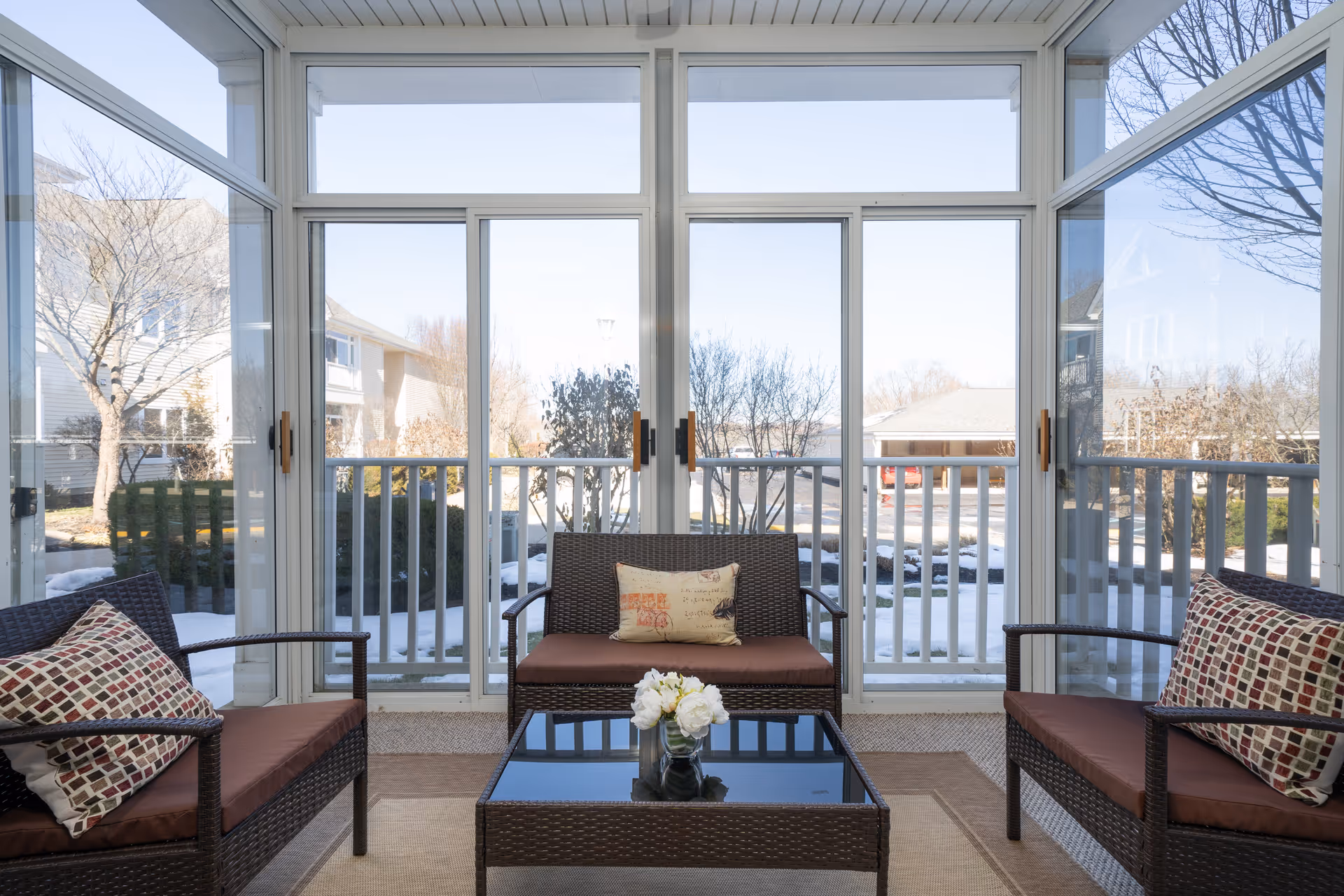 A bright enclosed porch area with large glass windows and sliding doors overlooking a snowy outdoor scene. The space features three dark wicker seating pieces with brown cushions and patterned pillows, arranged around a glass-top coffee table with a small vase of white flowers.
