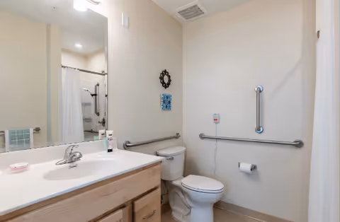 Bathroom with sink and mirror, toilet with grab bars and emergency pull cord, and a shower with curtain.