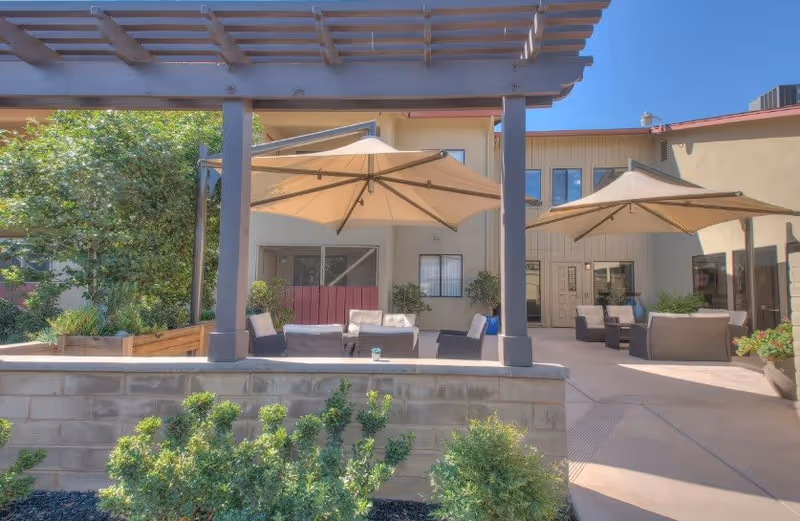 Outdoor patio area at Lodi Commons Senior Living with seating arrangements under large beige umbrellas, surrounded by plants and a pergola structure, adjacent to the building with windows and doors visible.