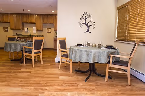 A dining area with two round tables covered with light blue tablecloths, each set with plates, bowls, and glasses. Wooden chairs with dark cushions surround the tables. The room has wooden flooring, a wall decoration shaped like a tree, and a window with wooden blinds. In the background, there is a kitchen area with wooden cabinets and countertops.