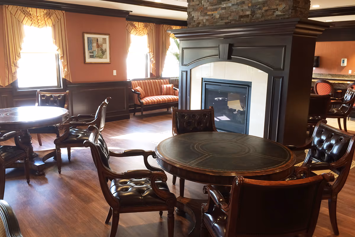 Cozy common room with round wooden tables, tufted leather chairs, a central fireplace, and a striped loveseat by sunlit windows.