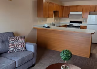 Interior view of a senior living facility showing a small kitchen with wooden cabinets, a white refrigerator, stove, and a countertop that extends into a breakfast bar. In the foreground, there is a gray upholstered armchair with a patterned cushion and a small round table with a decorative green plant.