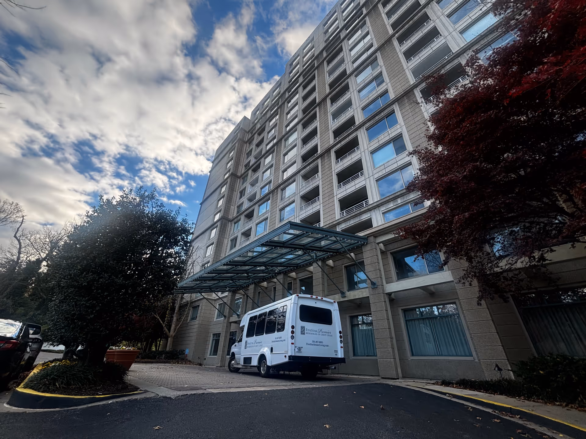 Exterior view of a multi-story senior living facility building with a covered entrance. A white shuttle bus with Five Star Premier Residences branding is parked under the entrance canopy. Trees and a partly cloudy sky are visible around the building.