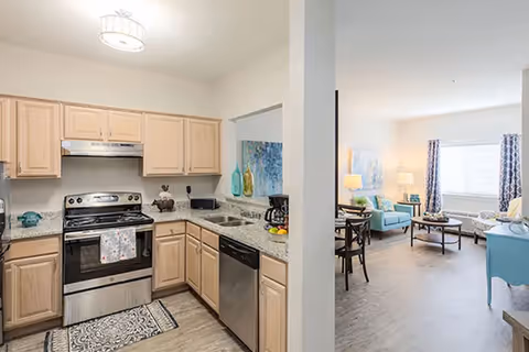 View of a kitchen with light wood cabinets, granite countertops, a stainless steel stove and dishwasher, and a coffee maker. Adjacent to the kitchen is a living room area with blue armchairs, a coffee table, a side table with a lamp, and large windows with curtains allowing natural light.