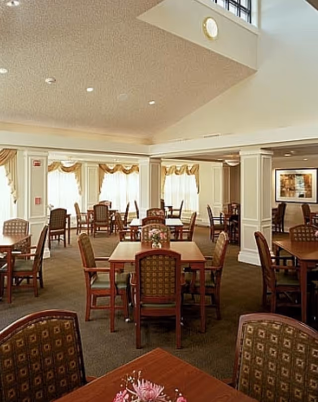 Dining room with wooden tables and upholstered chairs, large windows, and a high ceiling in a senior living facility.