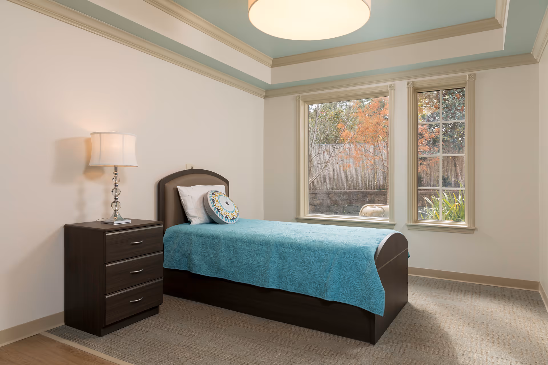 A simple, clean bedroom with a single bed covered in a turquoise quilt and a decorative round pillow. Next to the bed is a dark wooden nightstand with three drawers and a lamp with a white shade. The room has beige walls, a large window showing an outdoor view with trees, and a ceiling light fixture.