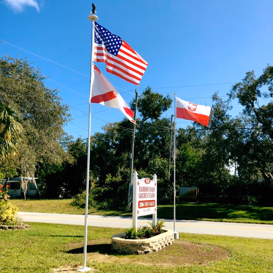 Outdoor scene showing three flagpoles with flags including the American flag, a red and white flag, and a red and white flag with an emblem. Below the flags is a sign that reads 'Harbor Oaks Golden Years' with a phone number and license information. The area is grassy with trees and a clear blue sky in the background.