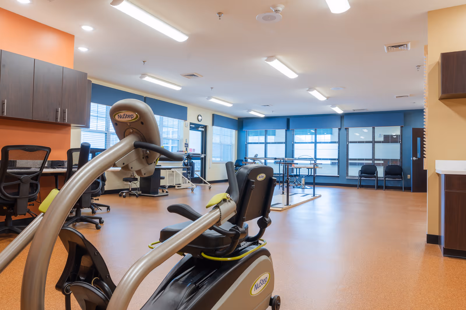 A spacious rehabilitation or physical therapy room with exercise equipment including a NuStep recumbent cross trainer in the foreground. The room has orange and beige walls, large windows with blue blinds, office chairs, and parallel bars for walking exercises. The floor is covered with a smooth, brown surface and the ceiling has multiple fluorescent lights.
