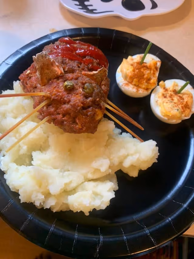 A black paper plate with mashed potatoes, a cat-shaped meatloaf topped with ketchup and peas and two deviled egg halves on the side.