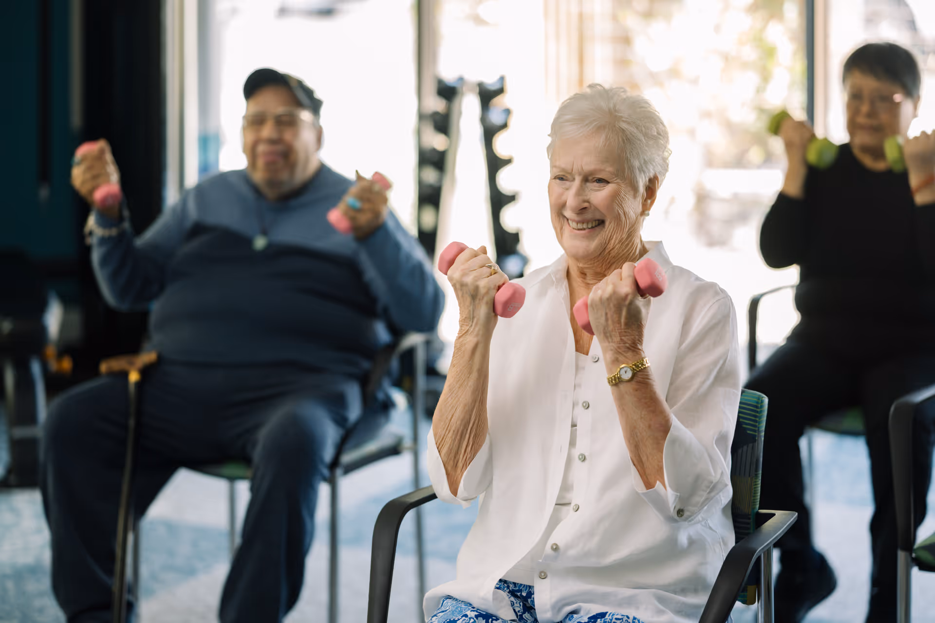 Three elderly individuals seated in chairs indoors, each holding small dumbbells and engaging in a light exercise or fitness activity. The woman in the foreground is smiling and wearing a white shirt, while the two people in the background are also lifting weights.