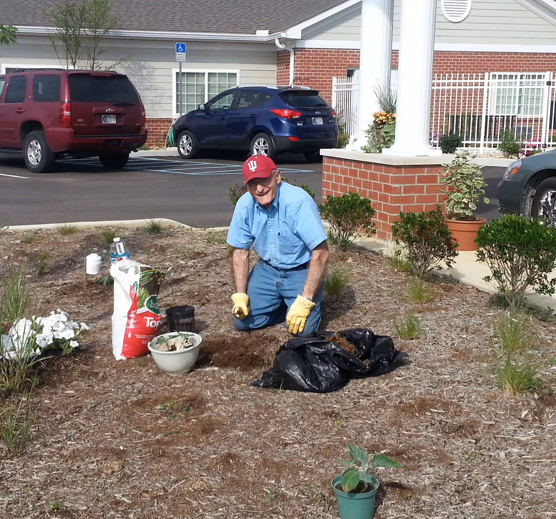 An elderly man wearing a red cap, blue shirt, jeans, and yellow gardening gloves is kneeling on the ground while gardening in a landscaped area outside a building. There are gardening supplies such as a bag of topsoil, a bowl, and a black plastic bag nearby. Several cars are parked in the background near a brick building with white columns.