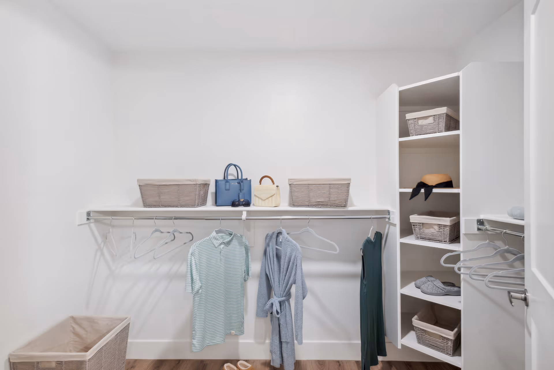A clean, organized walk-in closet with white walls and wooden floor. The closet features a hanging rod with a few empty hangers, a striped polo shirt, a blue robe, and a green dress hanging. On the top shelf, there are two wicker baskets and two handbags. To the right, there are built-in shelves holding wicker baskets, a hat with a black ribbon, and a pair of gray slippers. A large wicker basket is on the floor to the left.