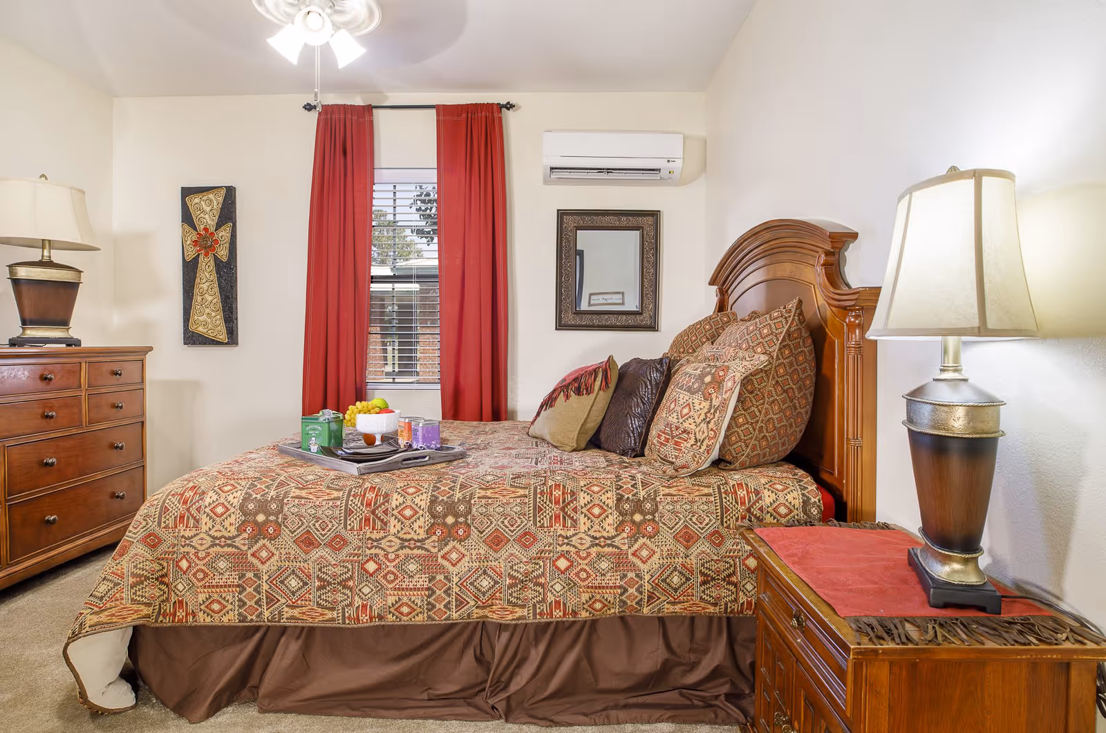 A cozy bedroom with a wooden bed frame and a patterned bedspread in shades of red, brown, and beige. The bed is adorned with multiple decorative pillows. A wooden nightstand with a lamp featuring a beige shade is on the right side of the bed. On the left side, there is a wooden dresser with a lamp and a decorative wall hanging above it. A window with red curtains and white blinds is centered on the back wall, and an air conditioning unit is mounted above the bed. A tray with snacks and drinks is placed on the bed.