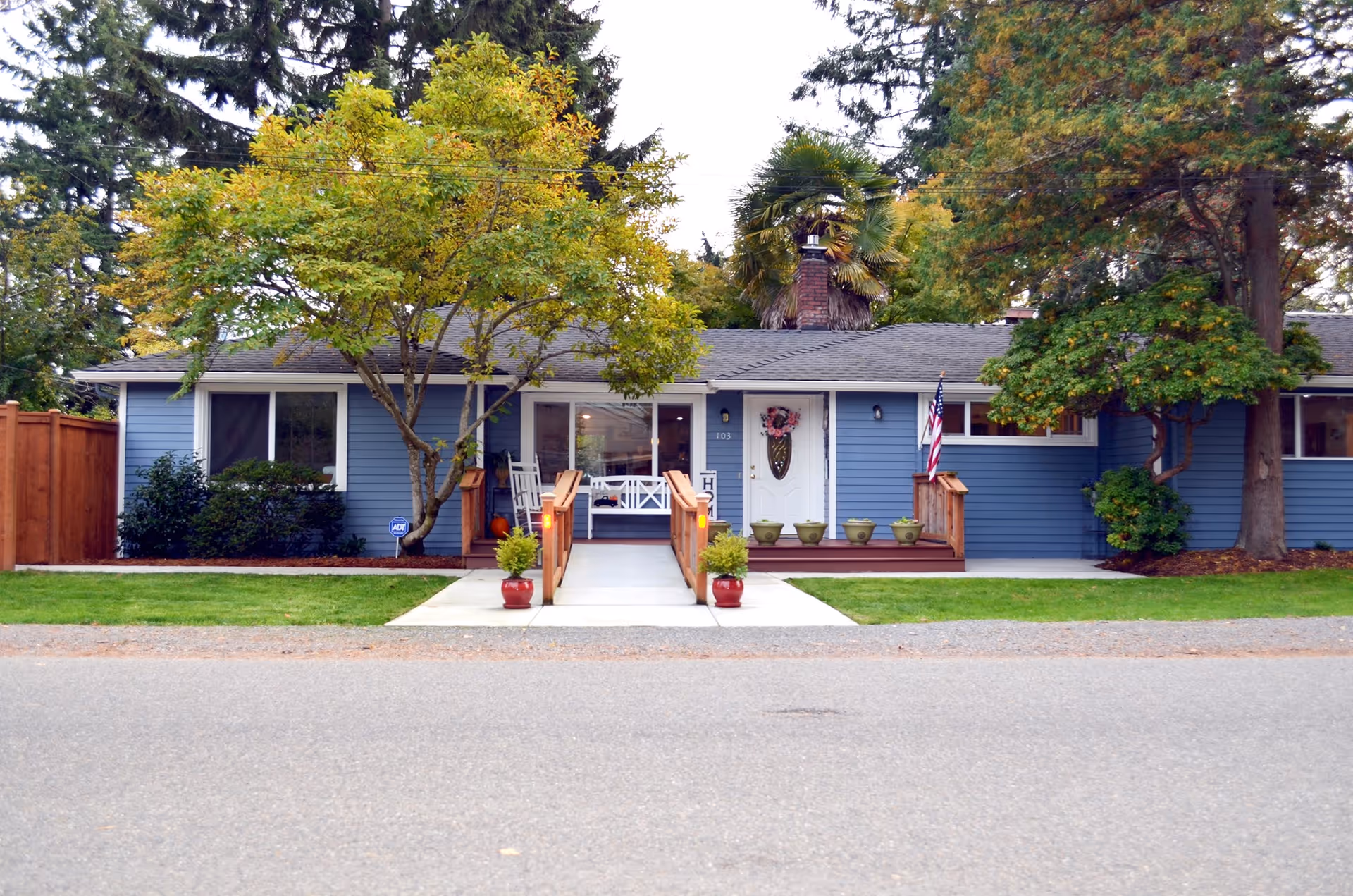 Front exterior of a single-story blue house with a ramped porch, potted plants, and surrounding trees.