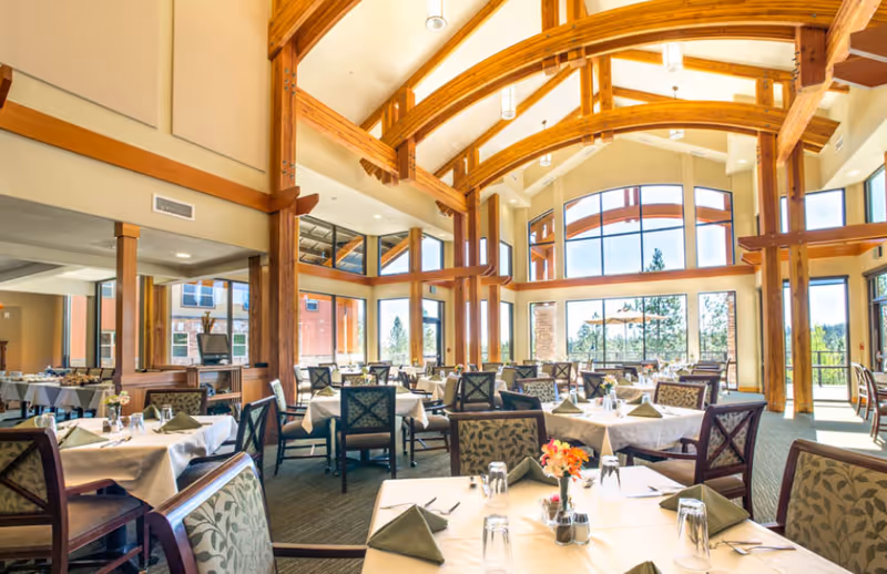 Bright dining room with vaulted wooden beams, tables set with white linens and large windows overlooking trees.