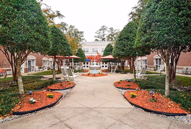 Outdoor courtyard area at Charter Senior Living of Newport News with neatly trimmed trees, flower beds with red mulch, a central fountain, and several white rocking chairs under red umbrellas. The courtyard is surrounded by a brick building with windows and doors.