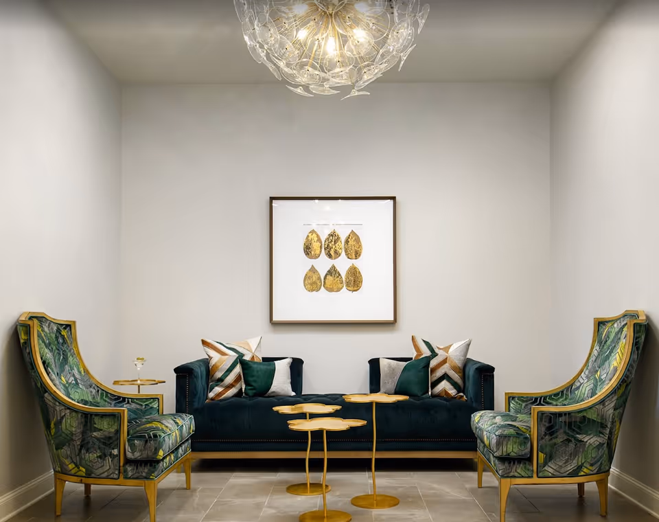 A modern living room with a dark green velvet sofa adorned with decorative pillows, two green patterned armchairs with wooden frames, three small gold side tables, a framed artwork featuring gold leaves on the wall, and a decorative chandelier hanging from the ceiling.