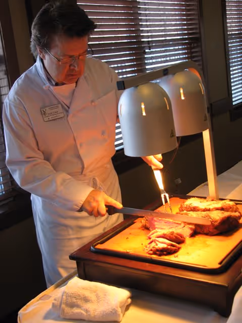 A chef wearing a white uniform and glasses is slicing cooked meat under two heat lamps on a carving station. The setting appears to be indoors with wooden blinds covering the windows in the background.