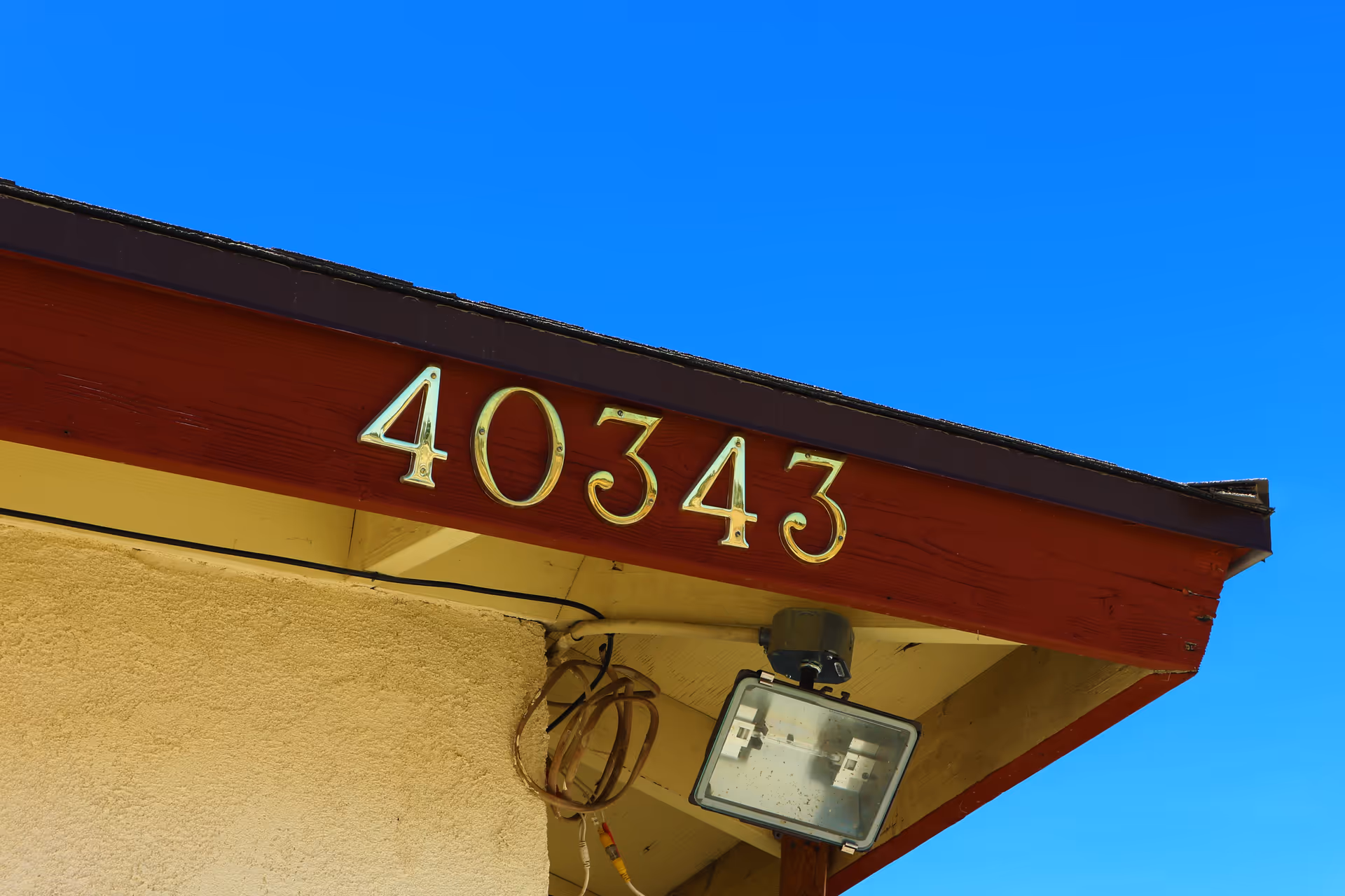 Close-up view of the corner of a building roof with the number 40343 displayed in gold-colored metal numbers on a red wooden fascia board against a clear blue sky. Below the numbers, there is a mounted outdoor floodlight and some coiled cables.