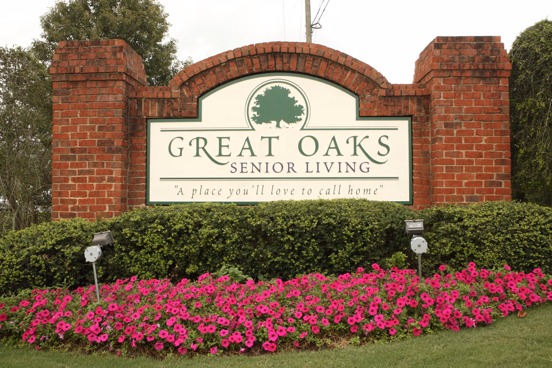 A large brick sign for Great Oaks Senior Living surrounded by green bushes and a bed of vibrant pink flowers, with a tree logo and the tagline 'A place you'll love to call home'.