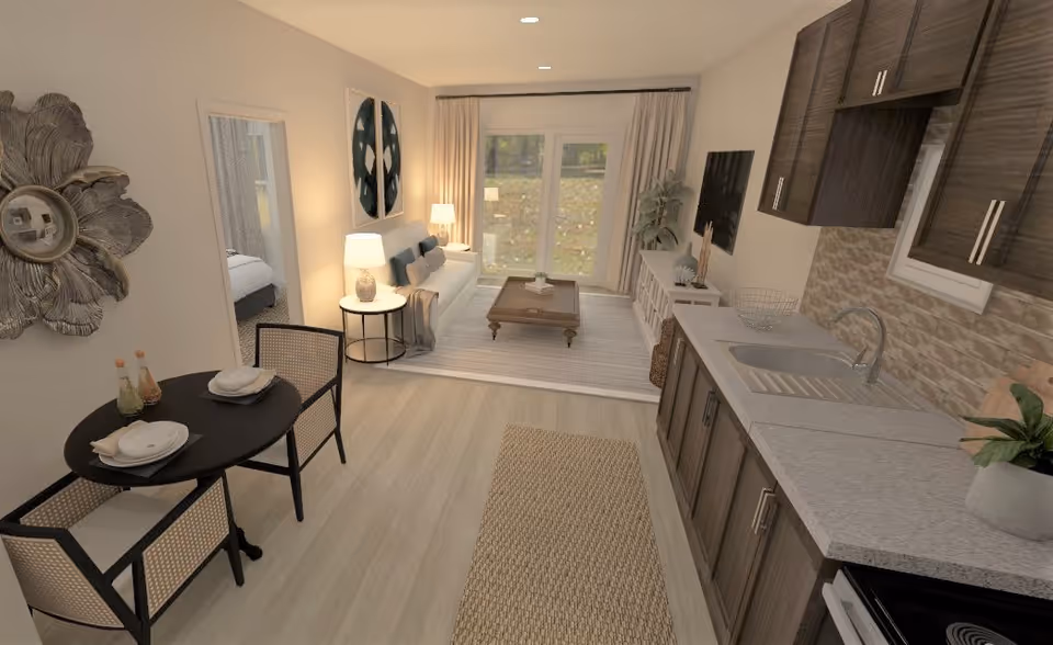 A modern open-concept living space featuring a kitchen with dark wood cabinets, a light countertop, and a sink on the right. On the left, there is a small dining area with a round table set for two and two chairs. Beyond the dining area is a living room with a white sofa, a wooden coffee table on a light rug, a side table with a lamp, and large glass doors with curtains leading outside. A bedroom is partially visible through an open door on the left.
