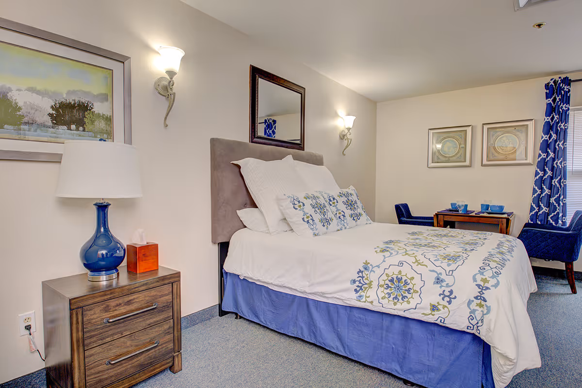 A neatly made bed with white and blue floral bedding in a senior living facility room. The room features a wooden nightstand with a blue lamp and a tissue box, wall-mounted light fixtures, a mirror above the bed, framed artwork on the walls, and a small dining table with two blue chairs near a window with blue patterned curtains.