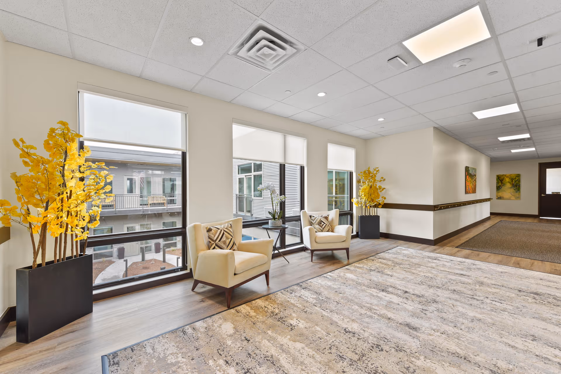 Bright senior living lounge area with two armchairs by large windows, yellow floral arrangements, and a wide rug in a hallway.