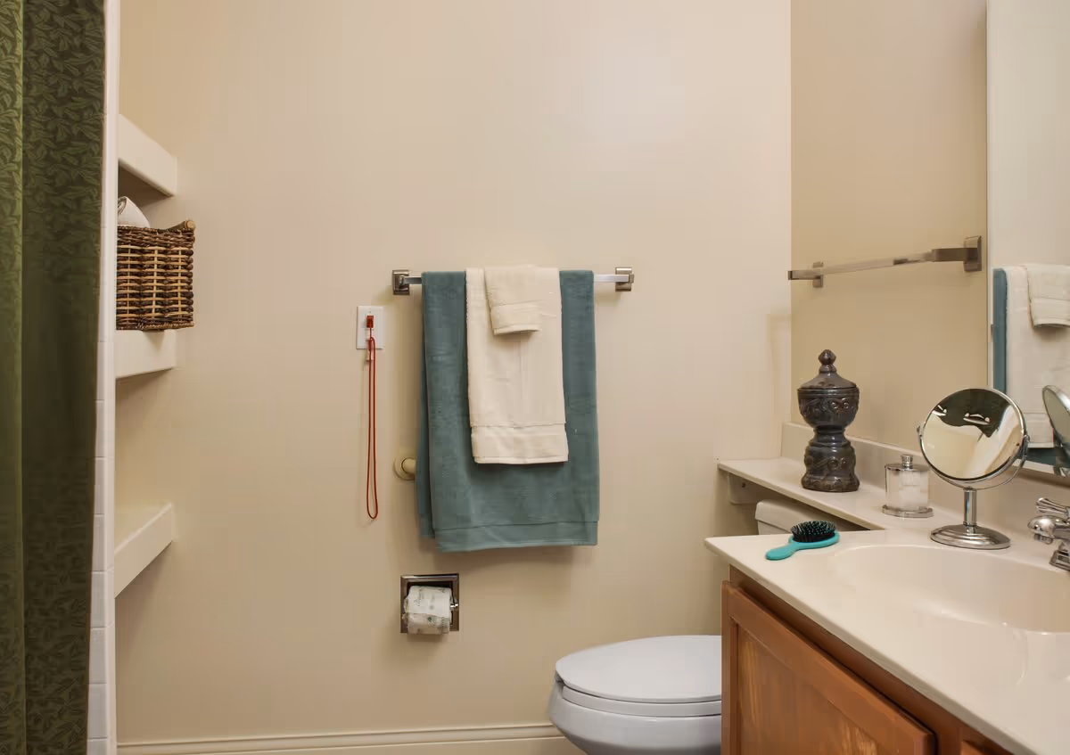 A bathroom with a toilet, a towel rack holding two towels, a countertop with a sink, a small round mirror, a hairbrush, a decorative container, and a glass jar. There is a green shower curtain partially visible on the left side and a wicker basket on a built-in shelf.