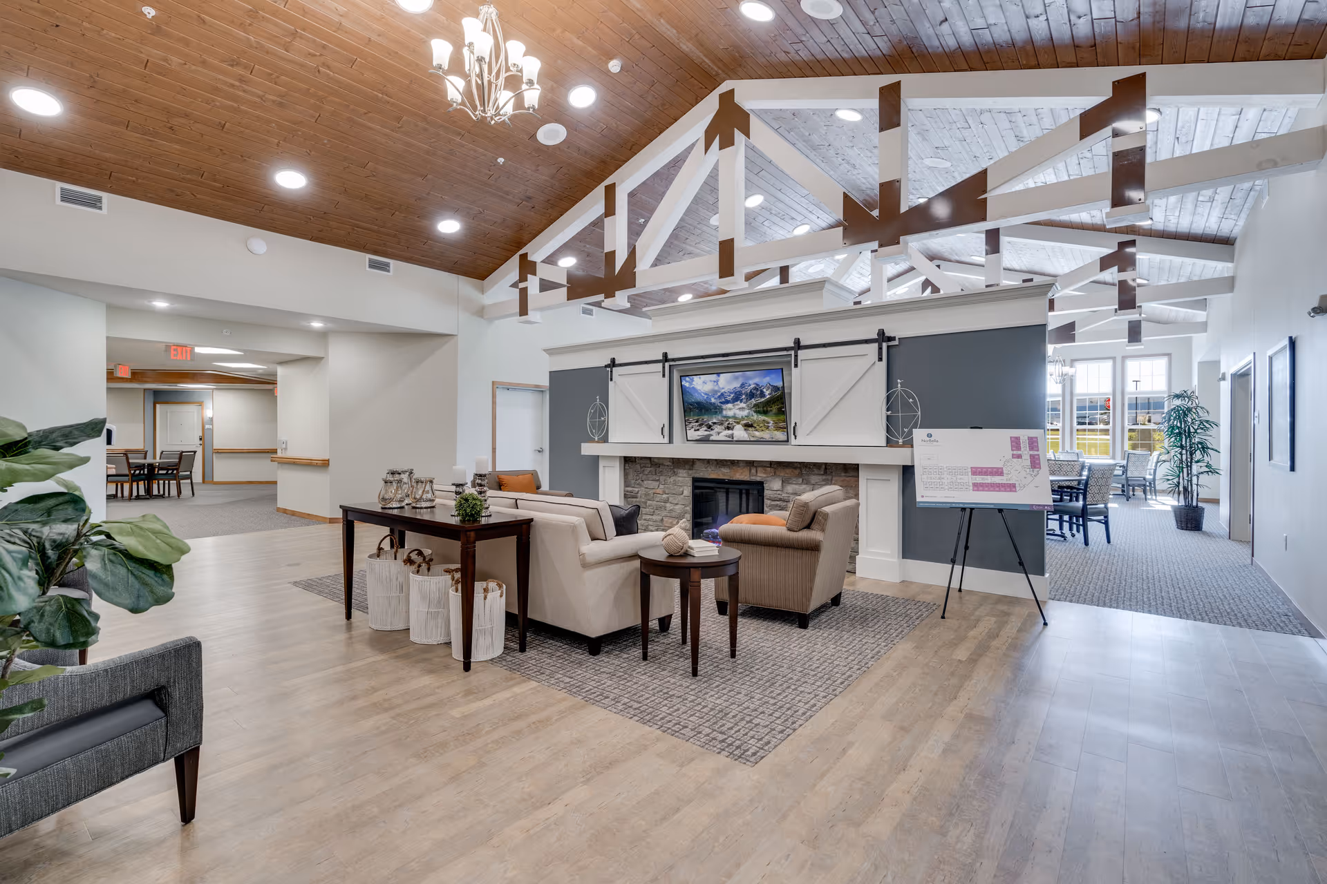 A spacious senior living facility common area with a high wooden ceiling featuring exposed beams and a chandelier. The room includes a seating area with beige and brown armchairs and sofas arranged around a stone fireplace with a mounted flat-screen TV above it. A wooden table with decorative items is placed behind the sofa. To the right, there is a floor plan on an easel and a dining area with tables and chairs near large windows letting in natural light. The flooring is a mix of wood and carpet, and there are plants adding greenery to the space.