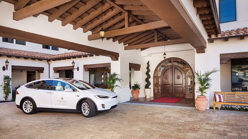Entrance of a senior living facility named Crestavilla featuring a covered driveway with a white car parked under the wooden beam canopy. The building has white walls, large wooden double doors with an arched window above, potted plants, and a wooden bench with cushions near the entrance.