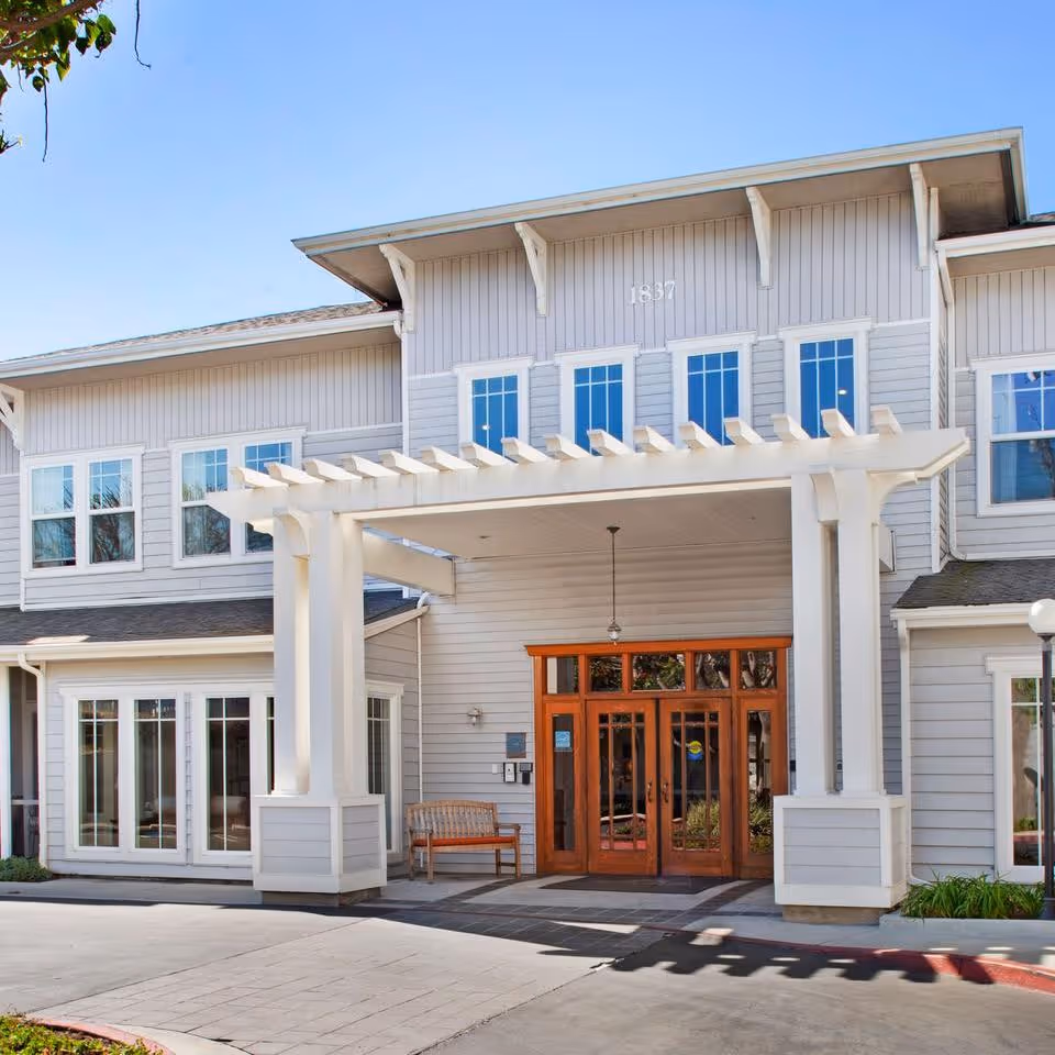 Front entrance of a light-gray two-story senior living building with a covered porte-cochere, wooden double doors, and multiple windows.