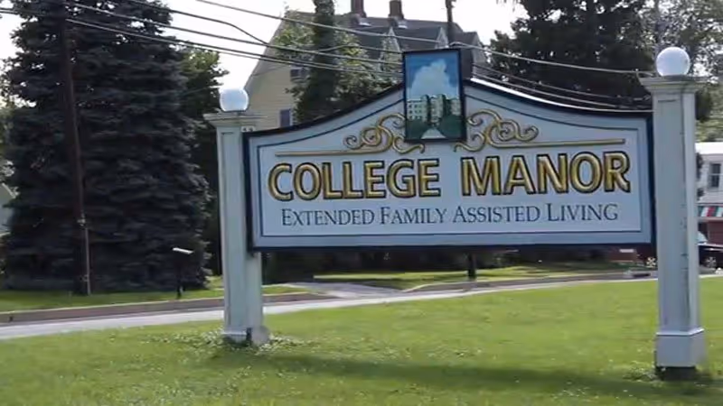 White decorative roadside sign reading "College Manor Extended Family Assisted Living" on a grassy lawn with trees and houses behind.