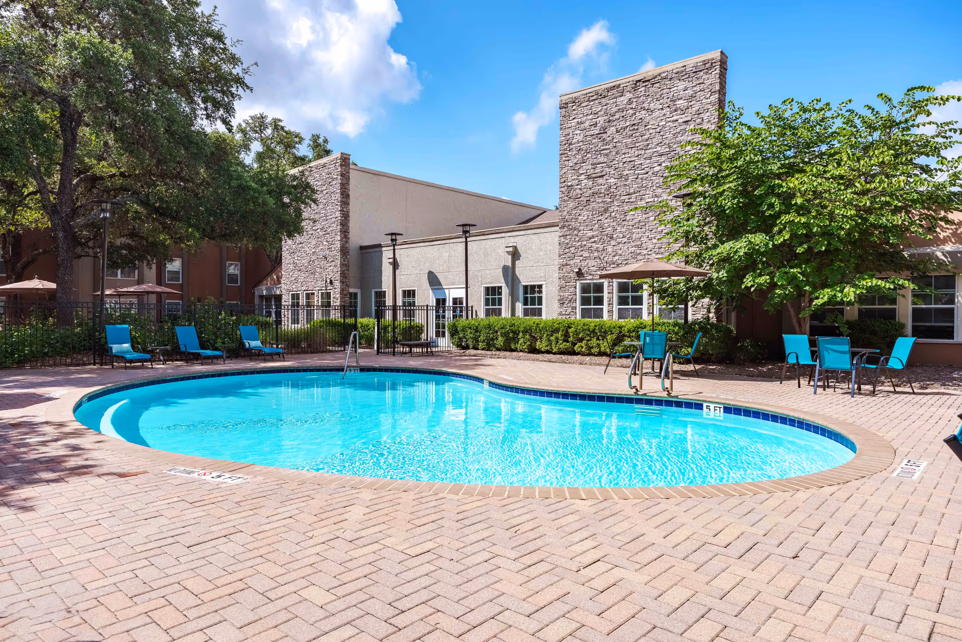 Outdoor swimming pool with clear blue water surrounded by a brick patio. Several turquoise lounge chairs and tables with umbrellas are arranged around the pool. The background shows a building with stone and beige walls, windows, and green trees under a blue sky with some clouds.