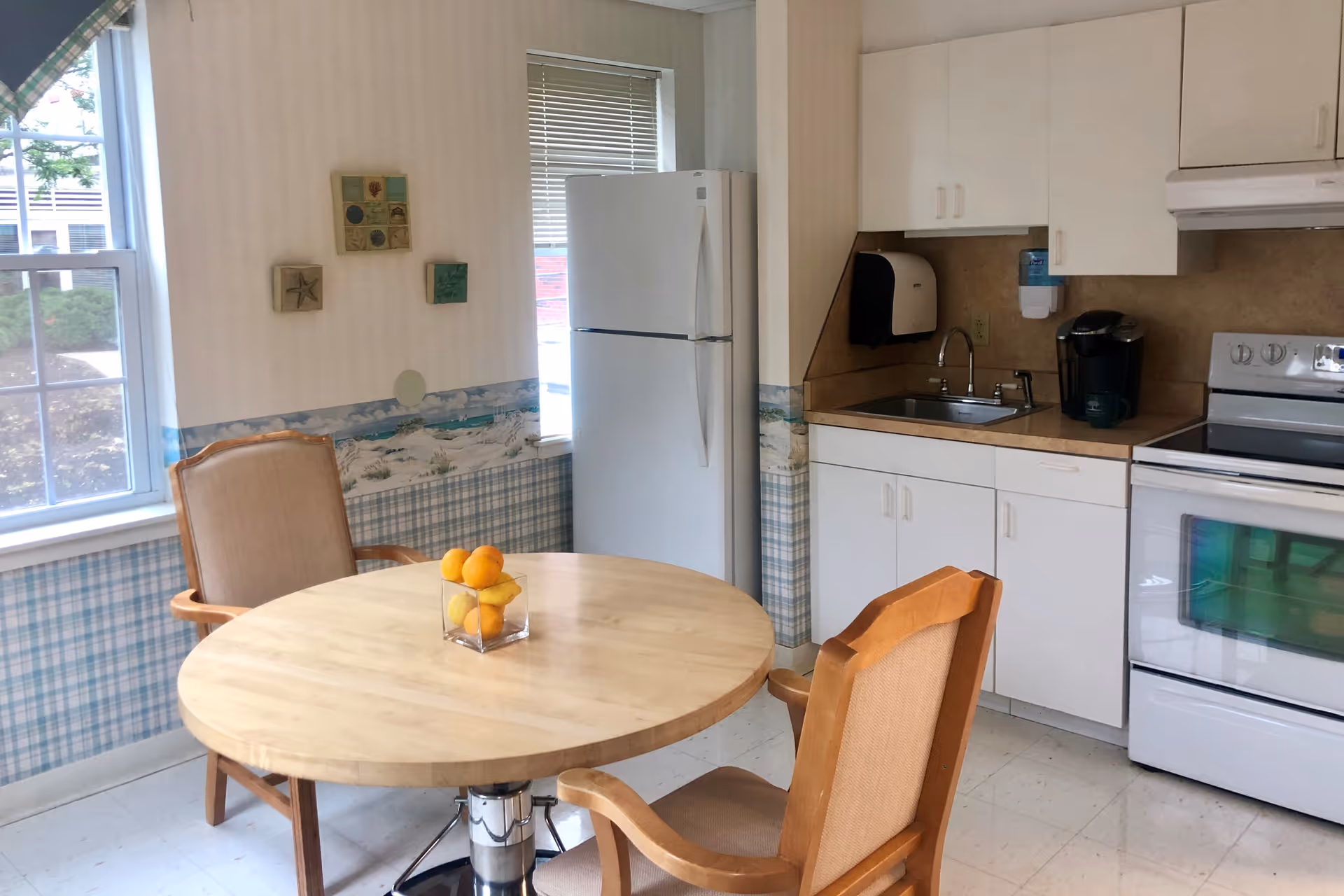 A small kitchen area with a round wooden table and two cushioned wooden chairs. On the table is a clear square vase holding oranges and lemons. The kitchen has white cabinets, a white refrigerator, a white stove, a sink with a faucet, a coffee maker, and a paper towel dispenser mounted on the wall. There is a window with blinds partially open, and the walls have a patterned wallpaper with a blue and white checkered lower half and a beach-themed border.