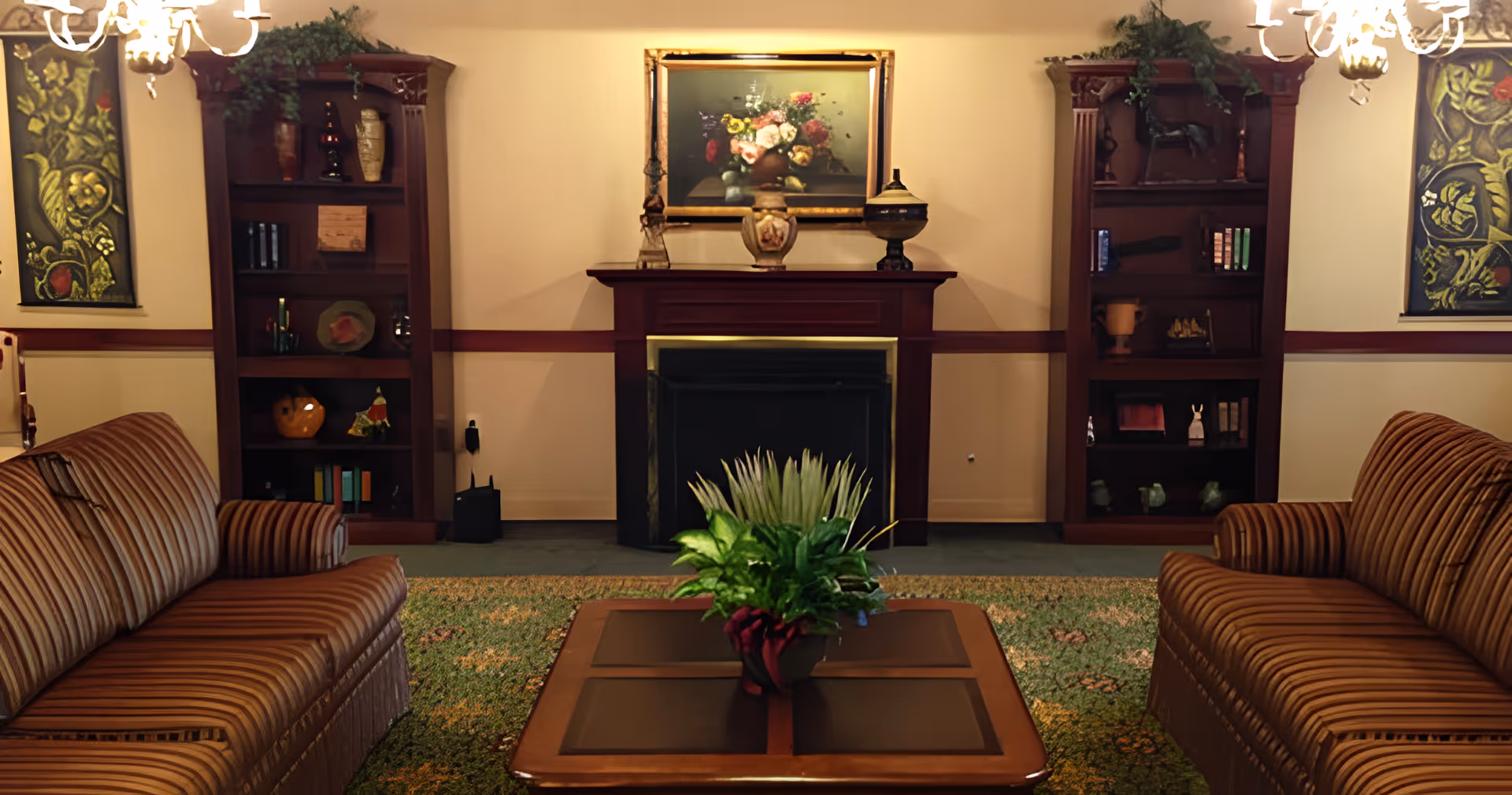 A cozy living room with two striped sofas facing each other across a wooden coffee table with a green plant centerpiece. Behind the table is a fireplace with a mantel decorated with vases and a framed floral painting above it. On either side of the fireplace are tall wooden bookshelves filled with decorative items and books. The room is warmly lit by chandeliers and has a patterned carpet.