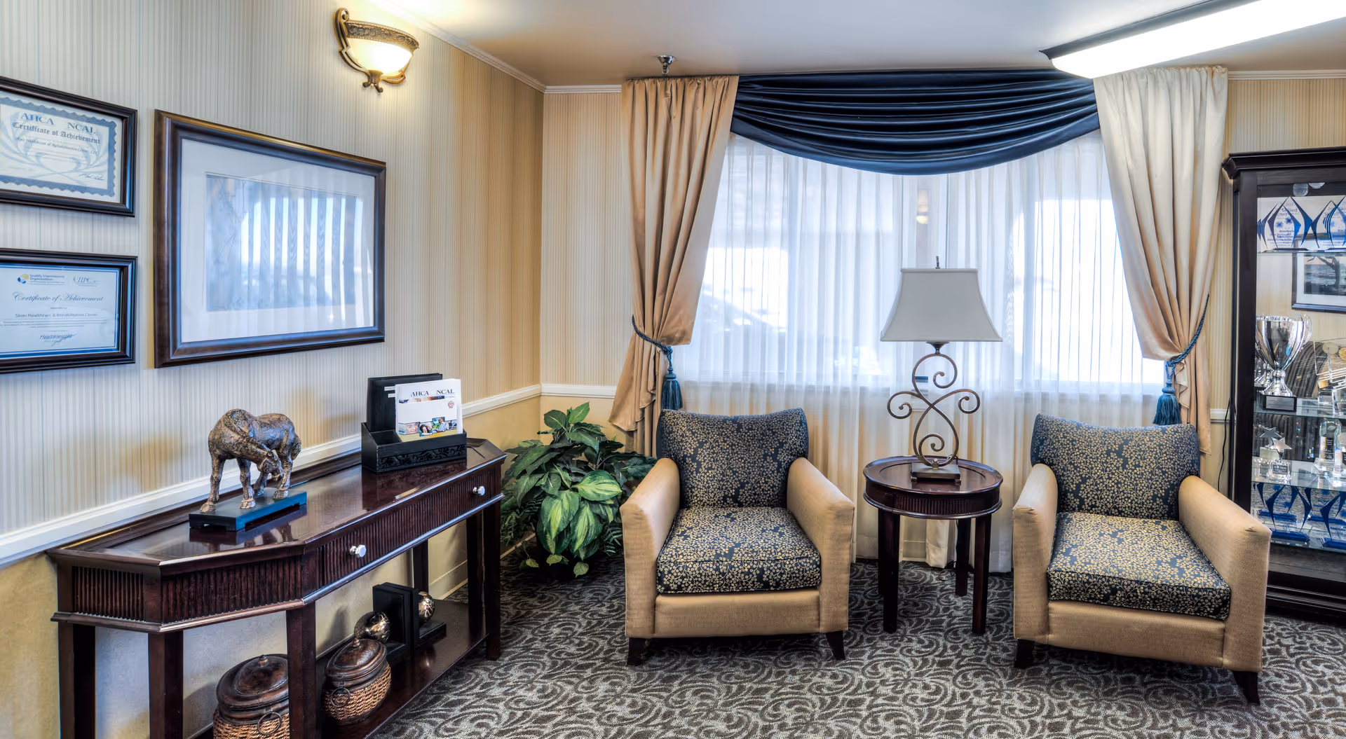 A cozy sitting area in a healthcare facility with two beige armchairs featuring patterned cushions, a small round wooden table with a decorative lamp between them, a large window with sheer white curtains and blue and beige drapes, a wooden console table with decorative items and framed certificates on the wall, and a glass display cabinet with trophies and awards.