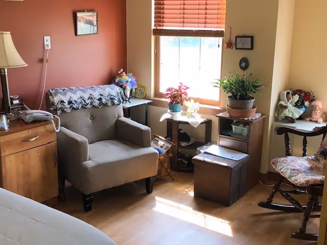 A cozy corner of a living room in an assisted living residence featuring a gray armchair with a crocheted blanket draped over the back, a wooden side table with a lamp and telephone, a window with closed blinds letting in sunlight, several potted plants on small tables, and a wooden rocking chair with floral cushions. The walls are painted in warm tones, and there are small decorative items and framed pictures on the walls and tables.