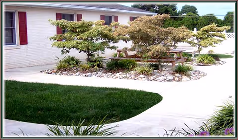 Outdoor courtyard area with a circular garden bed containing small trees and plants surrounded by a concrete walkway. A wooden picnic table is placed within the garden bed. The building exterior is visible with white walls and red window shutters.