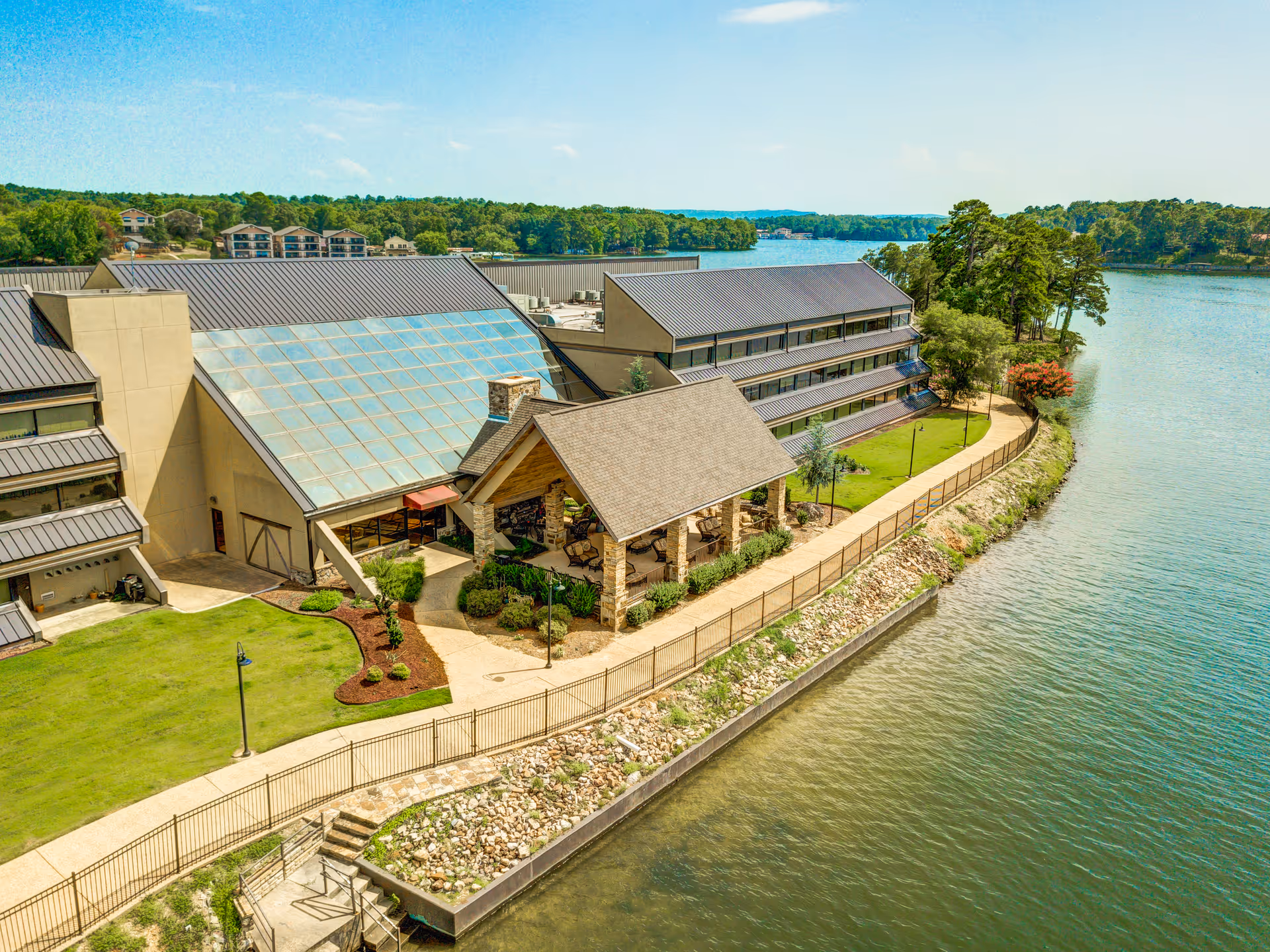 Aerial view of The Atrium at Serenity Pointe facility located beside a large body of water. The building features a large glass atrium, a covered outdoor seating area with stone pillars, and multiple floors with balconies. The surrounding area includes a well-maintained lawn, walking paths, and trees along the water's edge under a clear blue sky.
