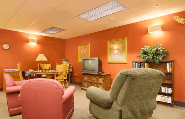A cozy living room with orange walls featuring two framed pictures, a wooden TV stand with an older television, a bookshelf with books and a plant on top, a green upholstered armchair, two red upholstered armchairs, and a wooden table with chairs and a lamp in the background.