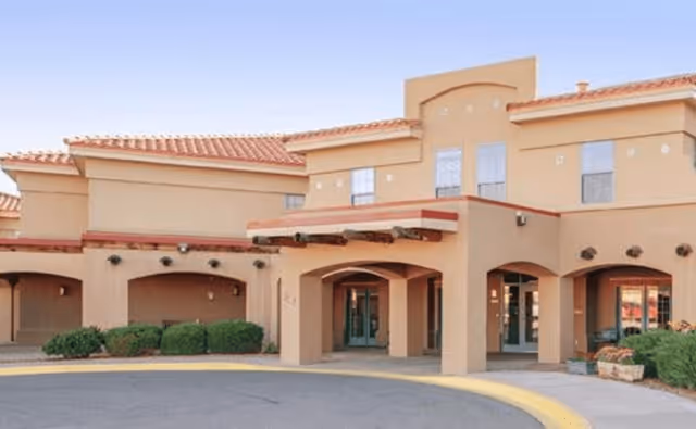 Exterior front view of a beige two-story building with a red-tiled roof and an arched entrance, surrounded by small bushes and a curved driveway.