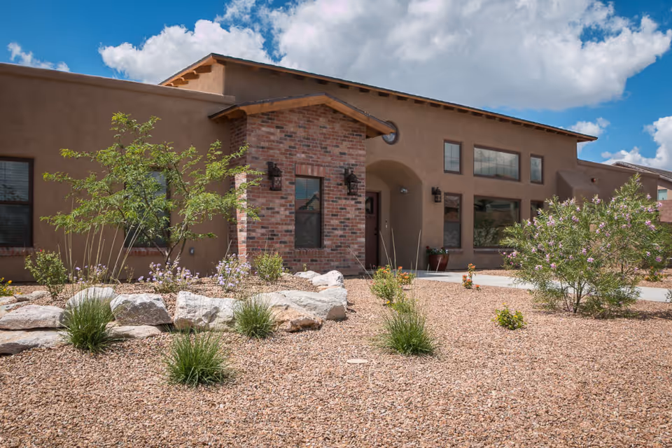 Single-story stucco and brick building with gravel landscaping, shrubs, and an arched entry under a blue sky.