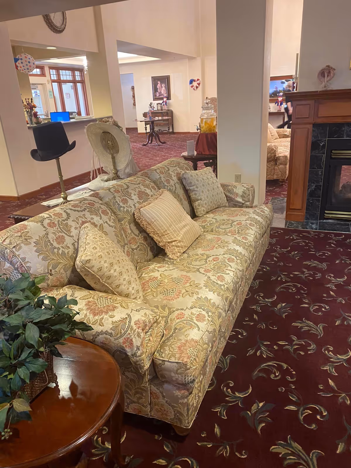 A cozy living room area with a floral patterned sofa adorned with multiple cushions. Next to the sofa is a wooden side table with a green leafy plant. In the background, there is a fireplace with a wooden mantle and a black tile surround. The room has a burgundy carpet with a floral design, and there are decorative items including hats on stands and framed pictures on the walls.