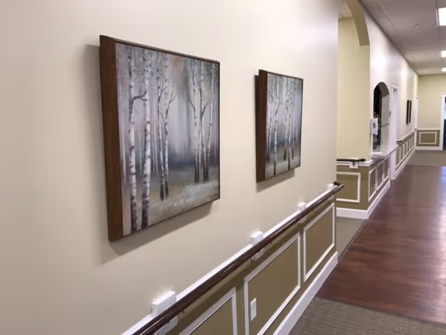 A hallway in a senior living facility with wooden handrails along the wall, two framed paintings of birch trees, and a polished wooden floor. The walls are painted beige with white trim and decorative molding. The hallway extends into the distance with an arched opening and additional wall decor.