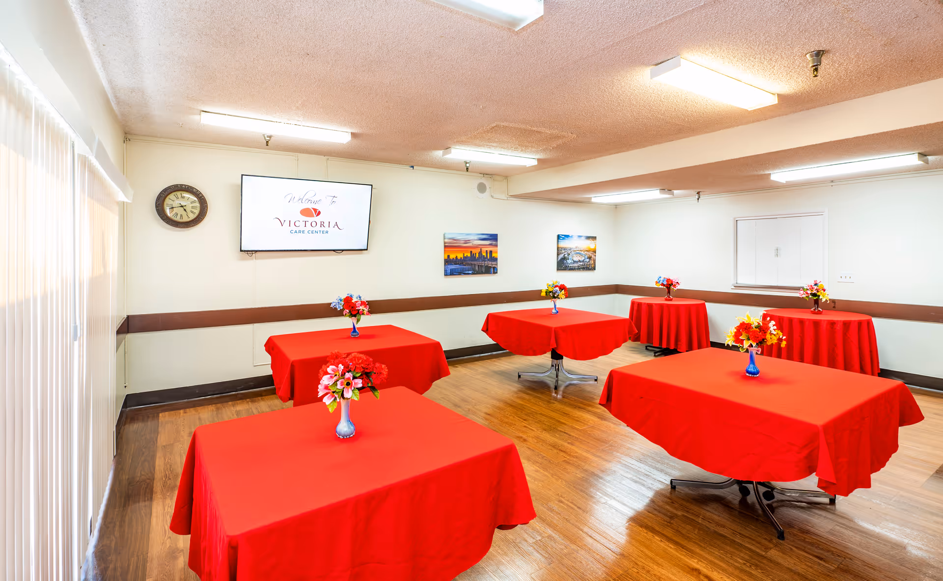 Room with several tables covered in red tablecloths and floral centerpieces, a wall-mounted TV displaying 'Victoria Care Center,' and wood flooring.