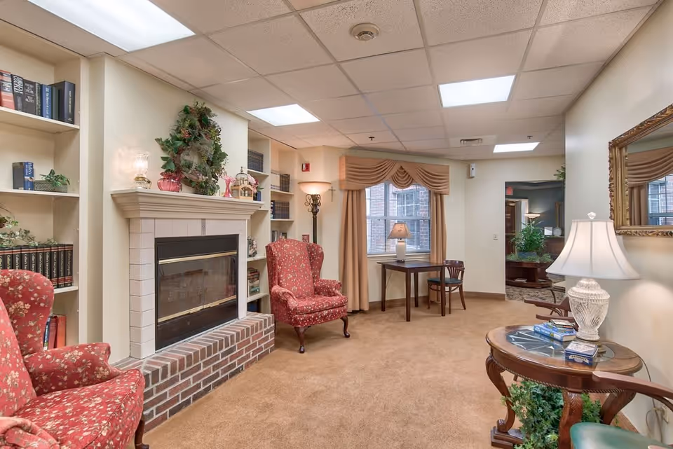 A cozy assisted living common area with two red floral armchairs placed near a white tiled fireplace with a brick base. Built-in bookshelves flank the fireplace, filled with books and decorative plants. A small wooden table with a lamp and chair is positioned in front of a window with beige curtains. A round wooden side table with a white lamp and books sits in the foreground, and a large mirror hangs on the wall. The room has beige carpet and a drop ceiling with fluorescent lighting.
