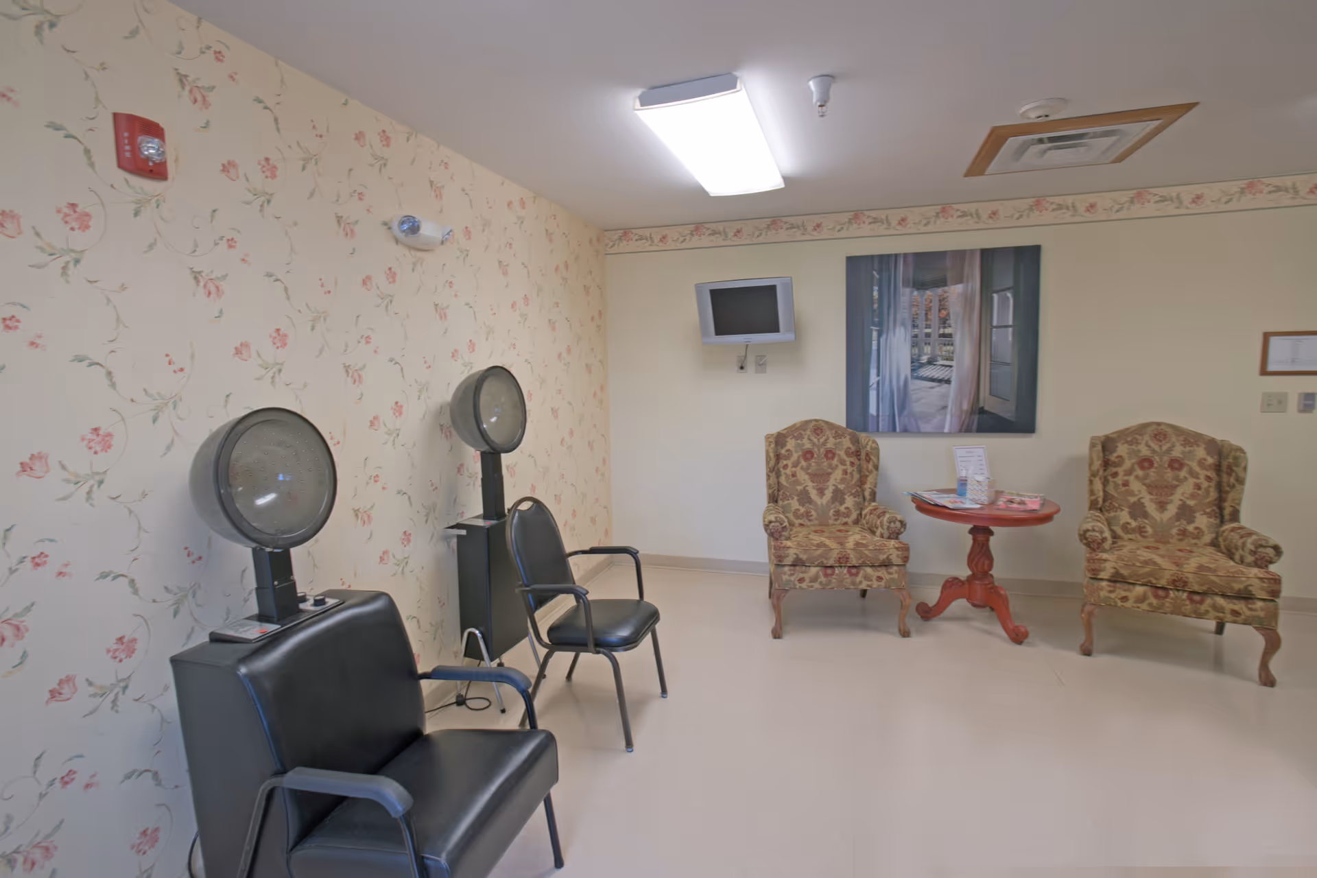 A small room with two vintage hair dryers and black chairs on the left side, and two patterned armchairs with a small round wooden table between them on the right side. The walls have floral wallpaper and a framed picture of a window with curtains. A small wall-mounted TV is also visible.